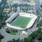 Progetto di riqualificazione dello Stadio Flaminio su un tavolo in primo piano, con lo stadio attuale sullo sfondo, a simboleggiare il futuro della casa della Lazio.