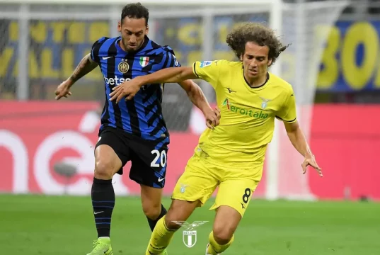 RASSEGNA STAMPA LAZIO: 40 Giorni al Via! Ecco le 5 Amichevoli per la Squadra di Sarri Un pallone da calcio sul campo di Formello con una sagoma di giocatori sullo sfondo che si allenano.
