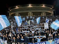 ✈️ Milan-Lazio, è Esodo: Oltre 2.000 Tifosi Biancocelesti Pronti a Invadere San Siro! Una curva gremita di tifosi della Lazio in trasferta allo stadio San Siro di Milano, che sventolano bandiere biancocelesti e sciarpe durante una partita notturna.