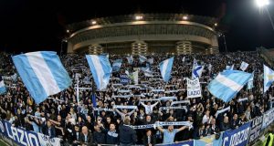 ✈️ Milan-Lazio, è Esodo: Oltre 2.000 Tifosi Biancocelesti Pronti a Invadere San Siro! Una curva gremita di tifosi della Lazio in trasferta allo stadio San Siro di Milano, che sventolano bandiere biancocelesti e sciarpe durante una partita notturna.