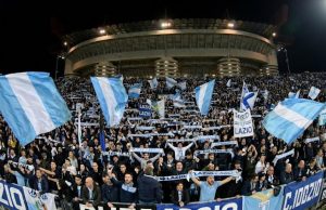 ✈️ Milan-Lazio, è Esodo: Oltre 2.000 Tifosi Biancocelesti Pronti a Invadere San Siro! Una curva gremita di tifosi della Lazio in trasferta allo stadio San Siro di Milano, che sventolano bandiere biancocelesti e sciarpe durante una partita notturna.