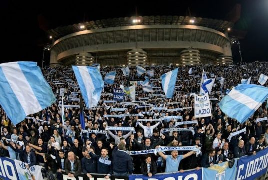 ✈️ Milan-Lazio, è Esodo: Oltre 2.000 Tifosi Biancocelesti Pronti a Invadere San Siro! Una curva gremita di tifosi della Lazio in trasferta allo stadio San Siro di Milano, che sventolano bandiere biancocelesti e sciarpe durante una partita notturna.