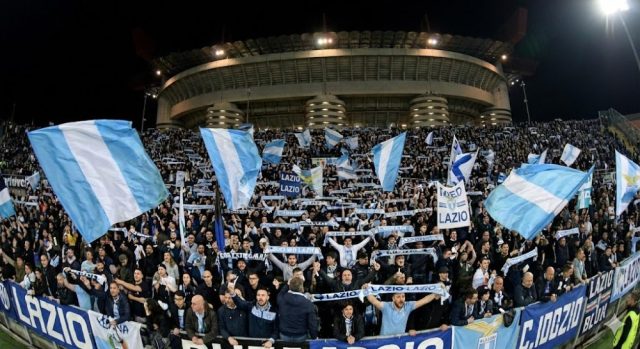 unnamed - 2025-11-27T163439.717 Una curva gremita di tifosi della Lazio in trasferta allo stadio San Siro di Milano, che sventolano bandiere biancocelesti e sciarpe durante una partita notturna.