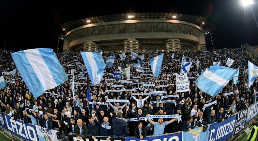 Una curva gremita di tifosi della Lazio in trasferta allo stadio San Siro di Milano, che sventolano bandiere biancocelesti e sciarpe durante una partita notturna.