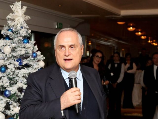 Claudio Lotito durante la cena di Natale della Lazio mentre parla del mercato bloccato e della situazione societaria. Claudio Lotito sorridente mentre parla ai microfoni, con una sovrimpressione grafica dei numeri finanziari (300M, 350M).